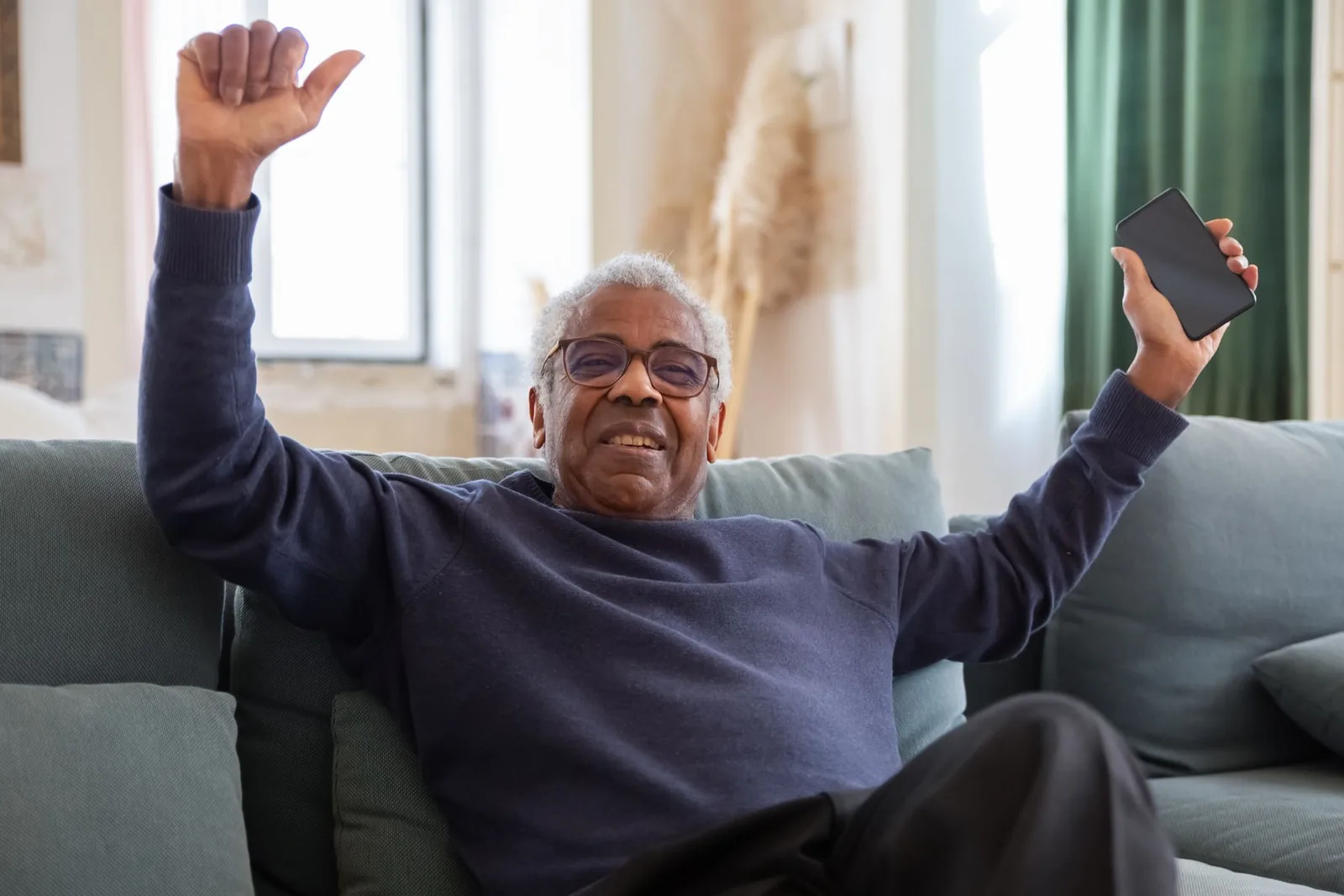 Homem idoso sorrindo com os braços erguidos no sofá de casa